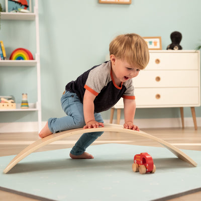 A young child balances on the TP Toys Active-Tots Pikler Style Climbing Triangle, Bridge, Climbing Cube & Balance Board—FSC® certified—while reaching for a red toy train on a light blue mat in the playroom.