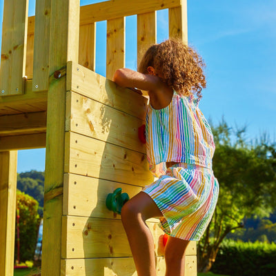 A child in a colorful striped outfit climbs the TP Toys Skywood Wooden Tower Climbing Frame with Ripple Slide (FSC® certified) outdoors on a sunny day.