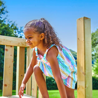 A young girl with curly hair, dressed in a colorful striped outfit, crouches on the TP Toys TP Skywood Mini Tower climbing frame (FSC® certified), enjoying the sunny day outdoors.