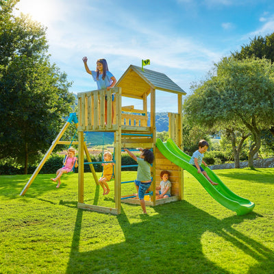 Five children play on a TP Toys Skywood Wooden Tower Climbing Frame with Ripple Slide, Sky Deck & Double Swing Arm (FSC® certified) in a grassy yard on a sunny day.