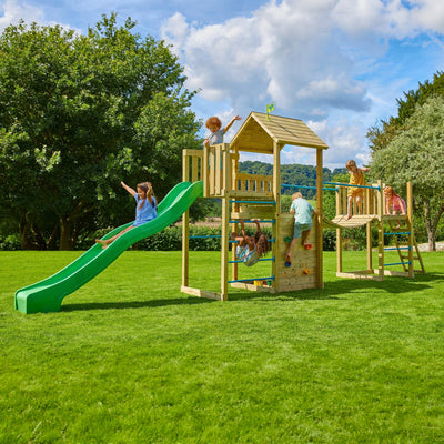 Children play on the TP Toys TP Skywood Wooden Tower Climbing Frame with Sky Deck, Super Wavy Slide, Sky Bridge & Mini Tower—an FSC® certified set—on a grassy lawn with trees and a blue sky in the background.