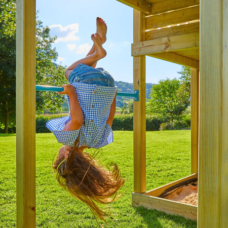 A child in a blue checkered dress hangs upside down from a horizontal bar on the TP Toys Skywood Wooden Tower Climbing Frame with Sky Deck, Super Wavy Slide, Sky Bridge & Mini Tower—FSC® certified—in a sunny grassy yard.
