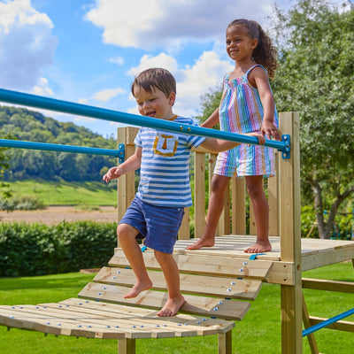 Two young children play outdoors on a sunny day with the TP Toys Skywood Wooden Tower Climbing Frame with Ripple Slide, Sky Bridge, Mini Tower & Double Swing Arm (FSC® certified), surrounded by green grass and trees.