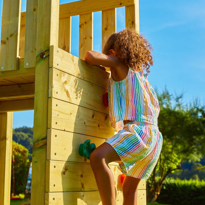 A young child climbs the TP Toys Skywood Wooden Tower Climbing Frame with Ripple Slide, Monkey Bars & Double Swing Arm (FSC® certified) on a sunny day.