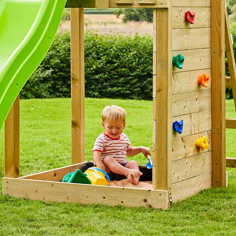 A young child plays with sand toys next to the TP Toys Skywood Wooden Tower Climbing Frame with Ripple Slide, Monkey Bars & Double Swing Arm (FSC® certified) on a grassy lawn.