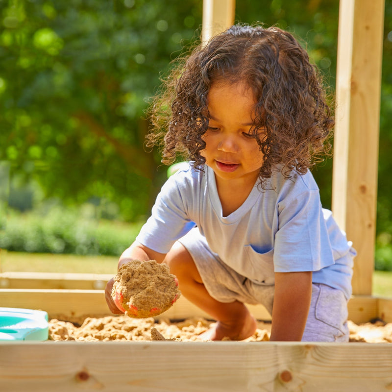 A young child with curly hair plays in a wooden sandbox next to the TP Toys Skywood Wooden Tower Climbing Frame with Ripple Slide, Monkey Bars & Double Swing Arm (FSC® certified), holding a handful of sand on a sunny day.