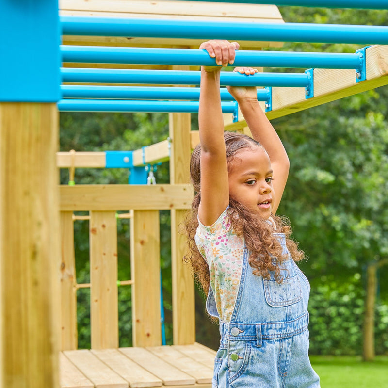 A young girl in denim overalls plays on the blue monkey bars of the TP Toys Skywood Wooden Tower Climbing Frame with Ripple Slide, Monkey Bars & Double Swing Arm – FSC® certified, at an outdoor playground.
