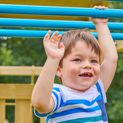 A young boy smiles as he hangs from blue Monkey Bars on the TP Toys Skywood Wooden Tower Climbing Frame with Ripple Slide, Monkey Bars & Double Swing Arm (FSC® certified), set in a playground with greenery in the background.