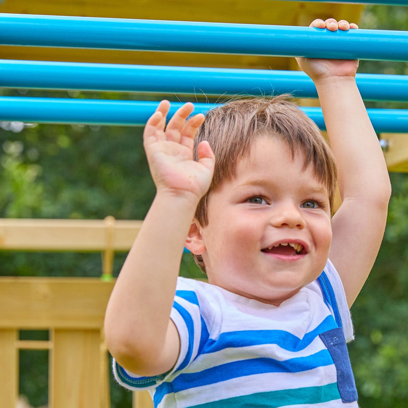A young boy smiles as he hangs from blue Monkey Bars on the TP Toys Skywood Wooden Tower Climbing Frame with Ripple Slide, Monkey Bars & Double Swing Arm (FSC® certified), set in a playground with greenery in the background.