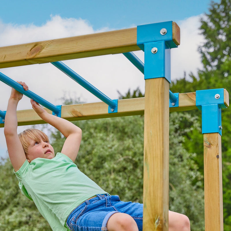 A young boy in a green shirt and denim shorts hangs from the monkey bars of the TP Toys Skywood Wooden Tower Climbing Frame with Ripple Slide, Monkey Bars & Double Swing Arm – FSC® certified, outdoors with trees and sky in the background.