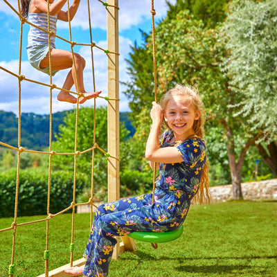 Two children play outside: one climbs a rope net on the TP Toys Skywood Wooden Tower Climbing Frame with Ripple Slide, Sky Deck, Skyline & Flying Fox Add On, while the other, in a floral jumpsuit, smiles from a swing amid trees and grass.