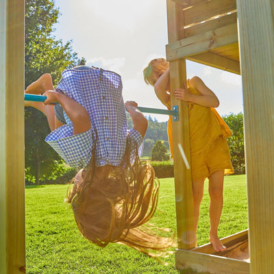 Two children play outside; one hangs upside down from a bar, while the other stands nearby on a TP Toys Skywood Wooden Tower Climbing Frame with Ripple Slide, Sky Deck, Skyline & Flying Fox. Sunlight filters over green grass and trees nearby.