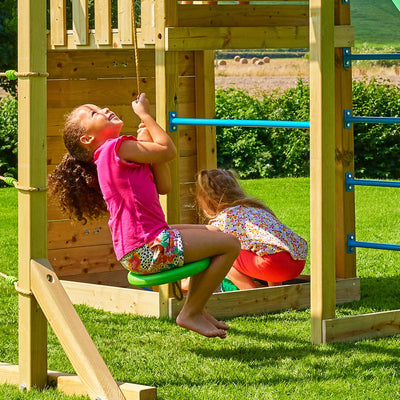 Two children enjoy the TP Toys Skywood Wooden Tower Climbing Frame with Ripple Slide, Sky Deck, Skyline & Flying Fox Add On—one swings on a rope seat while the other explores the modular play tower.