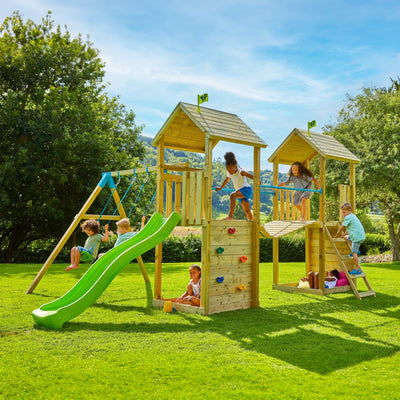 Children play on the TP Toys TP Skywood Wooden Tower Climbing Frame with Ripple Slide, Sky Bridge, Additional Play Tower & Double Swing Arm – FSC® certified, enjoying the set outdoors under a clear sky surrounded by trees.