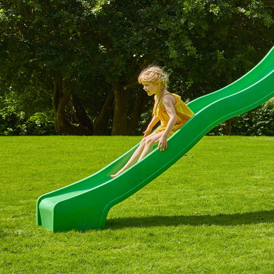 A young girl in a yellow dress slides down the super wavy slide from the TP Toys Skywood Wooden Tower Climbing Frame with Sky Deck, Monkey Bars, and Skyline with Rapide Swing Seat—FSC® certified—on a sunny day with grass and trees in the background.