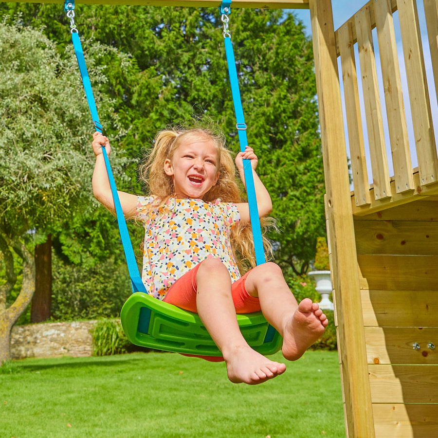 A young girl smiles as she swings on a green swing seat outdoors, with grass, trees, and the TP Toys Skywood Wooden Tower Climbing Frame featuring a super wavy slide, monkey bars, skyline, and extra play tower in the background.