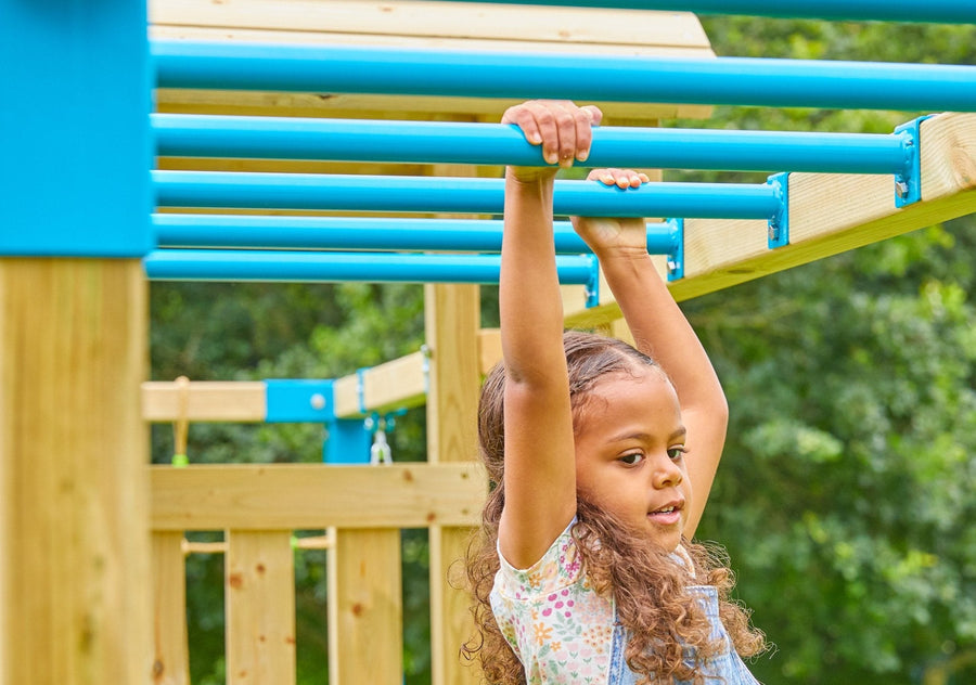 A young girl in denim overalls hangs from the monkey bars on the TP Toys Skywood Wooden Climbing Frame, featuring two extra play towers, slides, swings, and more—perfect for outdoor fun.