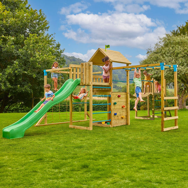 Six children play on the TP Skywood Wooden Tower Climbing Frame by TP Toys, featuring a Super Wavy Slide, Sky Deck, Monkey Bars, Skyline with Swing Seat & extra tower—FSC® certified—set on grass under a partly cloudy sky.