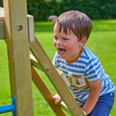 A young boy in a striped shirt climbs the TP Toys Skywood Wooden Climbing Frame, featuring two extra play towers and slides, smiling outdoors with green grass in the background.