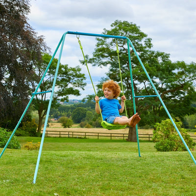 A young child with curly hair plays on a TP Toys Single Metal Swing Set in a grassy yard, surrounded by trees and a wooden fence under a cloudy sky.