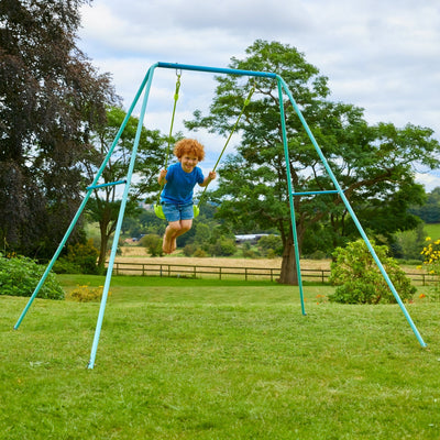 A young child with curly hair swings on the TP Toys TP Single Metal Swing Set with a braided rope seat in a grassy backyard, surrounded by trees and open fields.