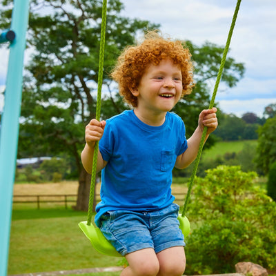 A young child with curly red hair sits on the green braided rope seat of the TP Single Metal Swing Set by TP Toys, outdoors among trees and grass.