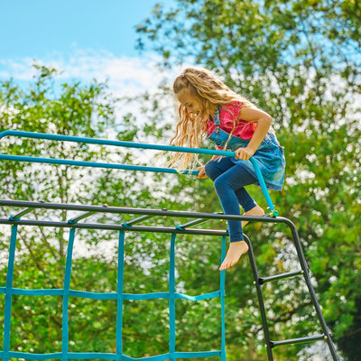 A young girl climbs on the TP Toys TP Explorer Metal Climbing Frame Black Edition with Ripple Slide, Swing Arm, Monkey Bridge & Basketball Hoop, enjoying outdoor fun among green trees and blue sky.