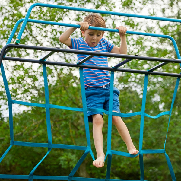 A young boy in a striped shirt and blue shorts climbs the TP Explorer Metal Climbing Frame Black Edition by TP Toys, gripping the top bars of this set that features a slide, swing arm, monkey bridge, and basketball hoop.