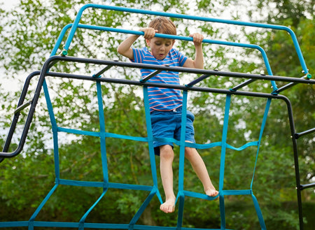 A young boy in a striped shirt and blue shorts climbs the TP Explorer Metal Climbing Frame Black Edition by TP Toys, gripping the top bars of this set that features a slide, swing arm, monkey bridge, and basketball hoop.