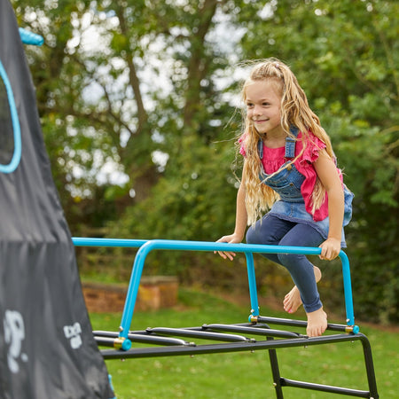 A young girl in a pink shirt and overalls climbs barefoot on the TP Toys Explorer Metal Climbing Frame Black Edition with Ripple Slide, Swing Arm, Jungle Run & Basketball Hoop outdoors, surrounded by trees and grass.