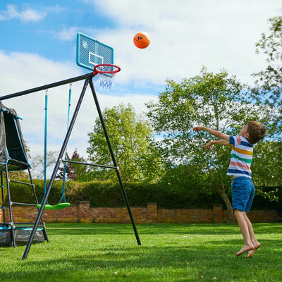 A young boy in shorts and a striped shirt throws a basketball toward the hoop on the TP Toys TP Explorer Metal Climbing Frame Black Edition with Ripple Slide, Swing Arm, and Monkey Bridge in a grassy backyard.
