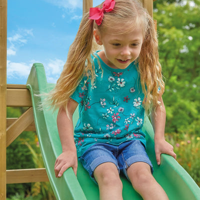A young girl with blonde pigtails and a pink bow plays on the TP Toys TP Robin Swing and 6ft Slide Set, sliding down the green wavy slide outdoors in a blue floral shirt and denim shorts.