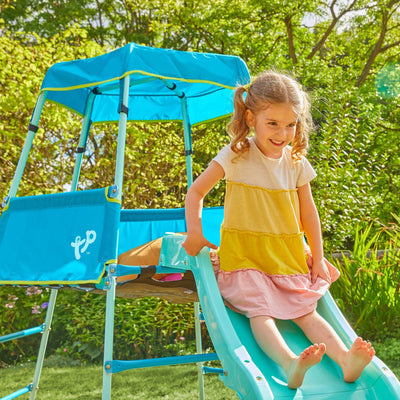 A young girl with pigtails slides down a blue slide attached to the TP Growable Adventurer Metal Climbing Frame by TP Toys in a sunny, green backyard.