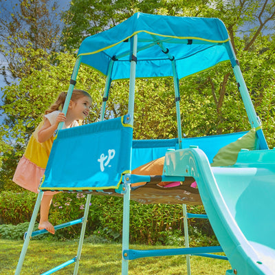 A young child climbs the TP Growable Adventurer Metal Climbing Frame by TP Toys, featuring blue and yellow colors, a canopy, and a slide, set in a grassy garden with trees in the background.