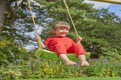 A young child in an orange outfit smiles while sitting on a roped Rapide seat outdoors, part of the TP Deluxe Double Metal Swing Set by TP Toys, featuring a powder-coated steel frame and surrounded by trees.