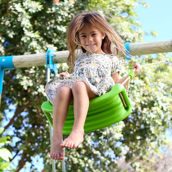 A young girl in a floral dress smiles as she sits on a TP Toys Sherwood Wooden Double Swing Set with 2 Roped Rapide Seats (PEFC certified), surrounded by trees and a blue sky.