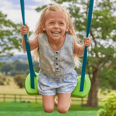 A young child with long blond hair smiles while swinging on the TP Toys TP Deluxe Single Metal Swing Set, featuring a green adjustable seat in a compact garden, surrounded by trees and grass outdoors.