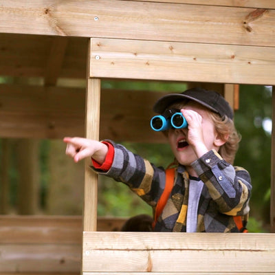 A boy in a cap and plaid shirt looks through blue binoculars and points outside while standing in the TP Toys TP Treetops Wooden Tower Playhouse with Toy Box – FSC® certified.