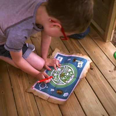 A child with red glasses uses a red marker to trace a path on a colorful treasure map while kneeling on the wooden floor of the TP Treetops Wooden Tower Playhouse with Toy Box by TP Toys.