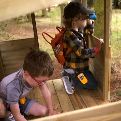Two boys play in the TP Toys Treetops Wooden Tower Playhouse with Toy Box—one kneels with binoculars and a backpack, while the other sits holding a walkie-talkie. The FSC® certified playhouse offers hours of imaginative outdoor fun.