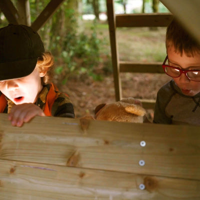 Two young children peer with surprise from inside the TP Treetops Wooden Tower Playhouse with Toy Box by TP Toys (FSC® certified), one in a cap and one in red glasses, as a plush toy peeks out between them.