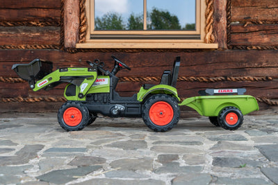 A TP Toys Falk Class Backhoe with Trailer, featuring a green and black design with red wheels, is parked on stone pavement in front of a wooden building with a window.