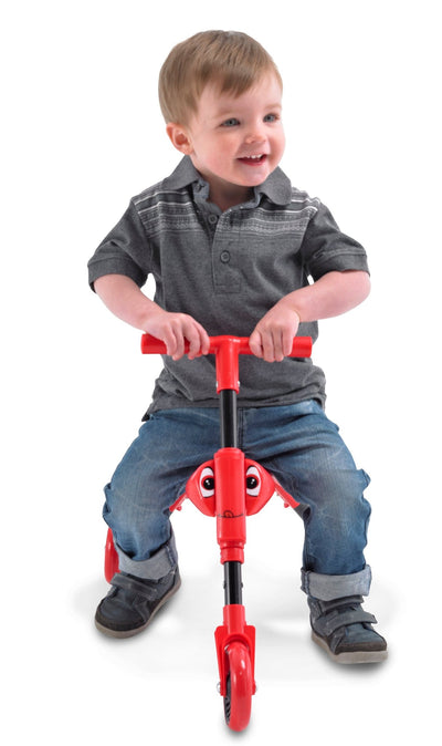 A young child in a gray shirt and jeans smiles while sitting on a red Scuttlebug Beetle Foldable Ride On Trike by Scuttlebug, looking slightly to the side against a white background.
