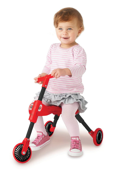 A young child in a pink striped shirt and gray skirt sits on a Scuttlebug Beetle Foldable Ride On Trike by Scuttlebug, which features black and red wheels, against a white background.