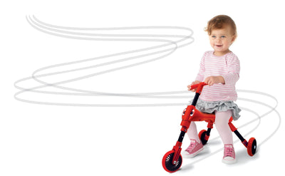 A young child in a pink and white outfit rides a small red Scuttlebug Beetle Foldable Ride On Trike by Scuttlebug, leaving curving grey lines on the white background.