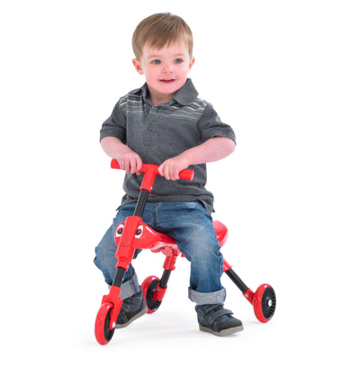 A young child in a grey shirt, blue jeans, and black shoes sits on a red Scuttlebug Beetle Foldable Ride On Trike by Scuttlebug, against a white background.
