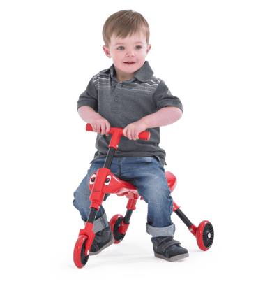 A young child smiles while sitting on a Scuttlebug Beetle Foldable Ride On Trike by Scuttlebug, holding the handlebars. The small red trike with black wheels is set against a plain white background.