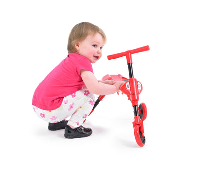 A toddler in a pink shirt and floral pants crouches beside a red Scuttlebug Beetle Foldable Ride On Trike by Scuttlebug, shown on a white background.