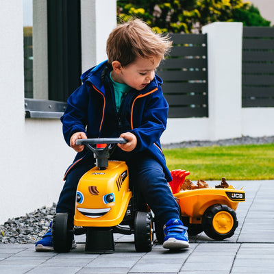 A young child in a blue jacket rides a TP Toys Falk Baby JCB Ride-On Tractor with Trailer, Rake & Shovel on a paved outdoor area near a lawn, looking down while sitting.