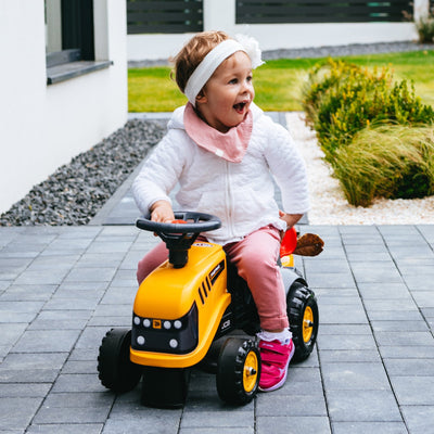 A toddler, wearing a white headband and pink pants, explores outdoors on the TP Toys Falk Baby JCB Ride-On Tractor with Trailer, Rake & Shovel.